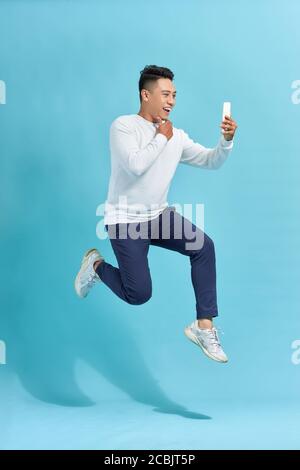 In voller Länge von gut aussehenden jungen Mann, der Telefon beim Springen Vor blauem Studiohintergrund Stockfoto