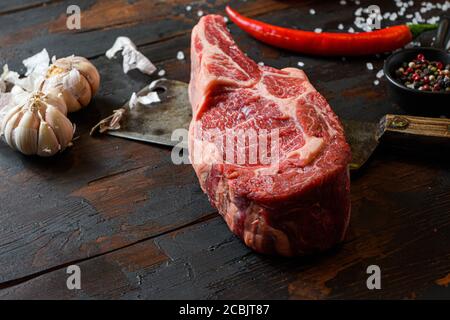 Rohes trockenes Cowboy Steak auf dunklem Holz Vintage-Tisch Hintergrund. Spencer Steak oder Beauty Steak BBQ mit Gewürzen, Rosmarin, Tomate, Chikie, Knoblauch Stockfoto