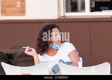 Huelva, Spanien - 13. August 2020: Reife Frau, die Gesichtsmaske trägt, um das Covid-19-Coronavirus zu vermeiden, raucht eine Zigarette auf einer Barterrasse Stockfoto
