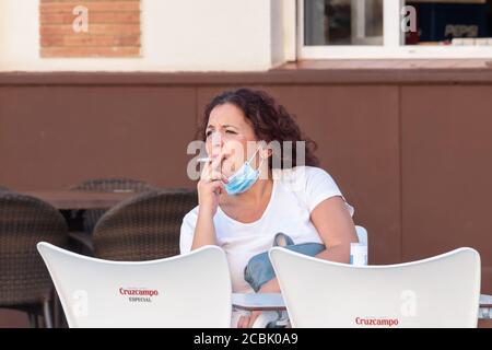 Huelva, Spanien - 13. August 2020: Reife Frau, die Gesichtsmaske trägt, um das Covid-19-Coronavirus zu vermeiden, raucht eine Zigarette auf einer Barterrasse Stockfoto