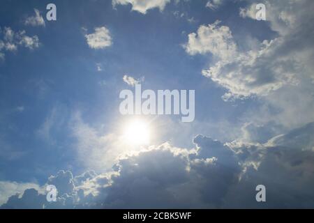 Blauer Himmel mit Wolken und Sonne im Zentrum. Die Sonne guckt hinter den Wolken hervor Stockfoto