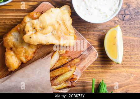 Detail von Fischchips mit Dip und Zitrone, pürierte Minzerbsen, Tartarsauce in Papierkegel auf Holz Schneidebrett Dip und Zitrone - gebratener Kabeljau, pommes frites Stockfoto