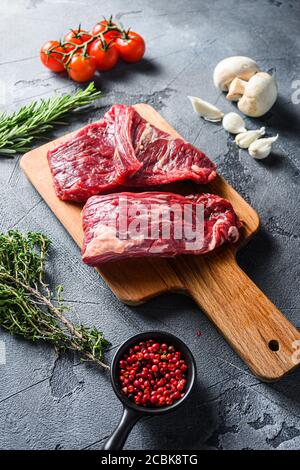 RAW Flap Steak Flanke geschnitten mit Machete, Rock Steak, auf Holz Schneidebrett, mit Kräutern Tomaten Pfefferkörner über grauen Stein Oberfläche Hintergrundseite Stockfoto