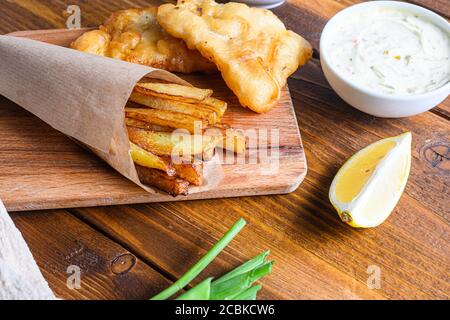 Detail von Fischchips mit Dip und Zitrone, pürierte Minzerbsen, Tartarsauce in Papierkegel auf Holz Schneidebrett Dip und Zitrone - gebratener Kabeljau, pommes frites Stockfoto