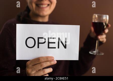 Lächelnde Frau mit OFFENEM Schild und Wein auf braunem Hintergrund. Wiedereröffnung lokaler Unternehmen Stockfoto