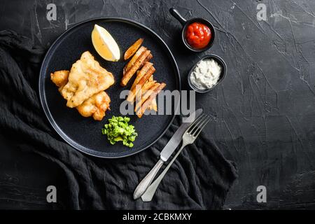 Fisch und Pommes. Traditionelles britisches warmes Gericht gebratener Fisch, Kartoffelchips, matschige Minzerbsen und Tartaresauce und Ketchup Blick von oben, schwarzer Stein Stockfoto