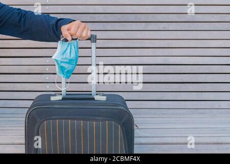 Hand hält Schutzmaske und Koffer. Handgriff von Gepäck und Einweg-medizinische Maske in der Hand. Bereit für die Reise während Coronavirus. Stockfoto