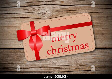 Geschenkkarte mit einem Text "Merry Christmas" und einem roten Band auf einem Holzhintergrund Stockfoto