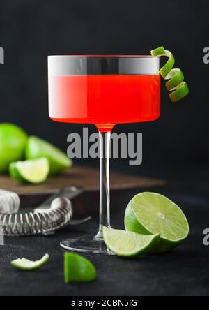 Kosmopolitischer Cocktail in modernem Kristallglas mit Limettenschale und frischen Limetten mit Sieb auf schwarzem Tischhintergrund. Stockfoto