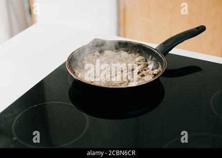 Gebratene Pilze und Zwiebeln auf Elektroherd in moderner weißer Küche. Prozess der Herstellung von Home Pizza, Zutaten aus der Nähe. Hausmannskost-Konzept Stockfoto