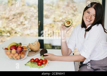 Junge glückliche Frau zeigt die Hälfte der perfekt reifen Avocado in der Hand und lächelt auf dem Hintergrund von frischem Obst und Gemüse in der modernen weißen Küche. Hea Stockfoto