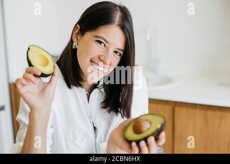 Junge glückliche Frau zeigt die Hälfte der perfekt reifen Avocado in den Händen und lächelt auf dem Hintergrund von frischem Obst und Gemüse in der modernen weißen Küche. Er Stockfoto