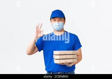 Kontaktlose Lieferung, covid-19 Versand, Virenschutzkonzept. Fröhliche asiatische männliche Kurier in blauer Uniform, medizinische Maske und Handschuhe, bringen Pizza Stockfoto