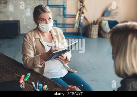 Ältere kaukasische Frau, die eine medizinische Maske trägt, hat eine Treffen mit einem Kunden zu Hause schriftlich etwas in der Notizbuch Stockfoto