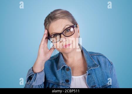 Ältere kaukasische blonde Frau mit Brillen berühren ihren Kopf und Blick auf die Kamera in einer blauen Jeansjacke auf einem studiowand Stockfoto
