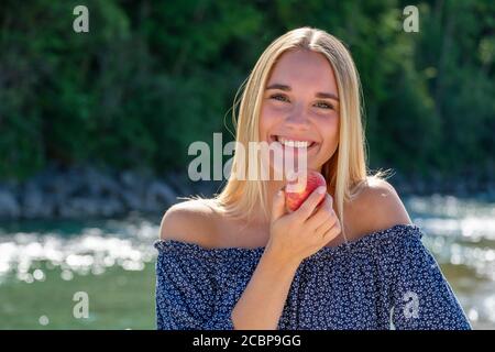 Porträt, junge blonde Frau lachend am Wasser, essen einen Apfel, Freude, Sommer, Isar, Bayern, Deutschland Stockfoto