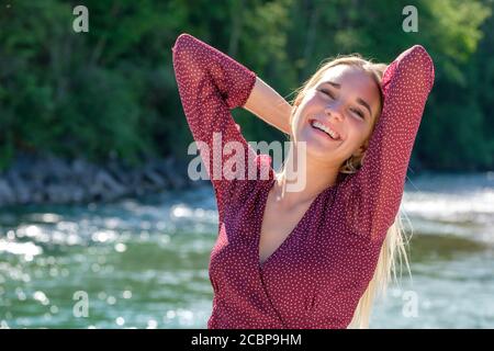 Portrait junge blonde Frau am Wasser, Arme hinter dem Kopf gekreuzt, glücklich, Freude, Sommer, Isar, Bayern, Deutschland Stockfoto