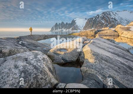 Person mit gelber Jacke Spaziergänge entlang der felsigen Strand von Tungeneset, Spiegelung der schneebedeckten Felsen Gipfel Devils Zähne, Devil's Zähne Stockfoto