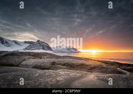 Felsiger Strand von Tungeneset, schneebedeckte Berggipfel im warmen Sonnenlicht über dem Meer, Steinfjorde, Senja Island, Skaland, Troms, Norwegen Stockfoto