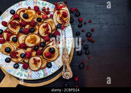 Mini Pfannkuchen mit Beeren auf einem gemusterten Teller mit silbernem Löffel im rustikalen Stil. Traditionelles Frühstück auf schwarzem Tisch Stockfoto