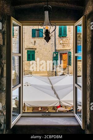Blick durch ein offenes Fenster einer Fassade eines alten Steinhauses mit hölzernen Fensterläden auf einer malerischen Straße der mittelalterlichen Altstadt Kotor, Montenegro Stockfoto