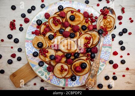 Mini Pfannkuchen mit Beeren auf einem gemusterten Teller mit silbernem Löffel im rustikalen Stil. Traditionelles Frühstück auf weißem Holz Stockfoto