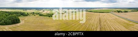 Luftweiten Panoramablick. Ländliche Landschaft mit Landstraße zwischen gelben Weizenfeldern, Dorf und Wald am Horizont unter bewölktem Himmel Stockfoto