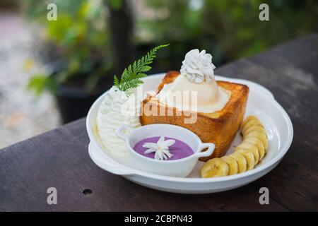 Vanilla Ice-Cream Honey Toast mit lila Kartoffelcreme serviert mit Banane in weißen Keramikplatte auf dem Holztisch. Stockfoto