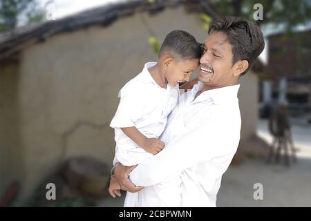 Glücklich ländlichen indischen jungen Vater und kleinen Sohn, die einander angucken und lachen, umarmen, beide tragen weiße traditionelle Tücher mit Dorf backgro Stockfoto