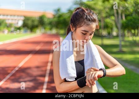 Schöne junge asiatische Frau stoppt laufen, um ihre Zeit zu überprüfen Auf ihrer smarten Uhr während ihres morgendlichen Laufs an Outdoor-Laufstrecke auf einer hellen sonnigen da Stockfoto