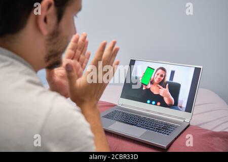 Junger Mann klatscht und spricht mit einer jungen Frau, die ihm am Telefon etwas zeigt. Junges Paar Online-Anruf-Konzept. Stockfoto