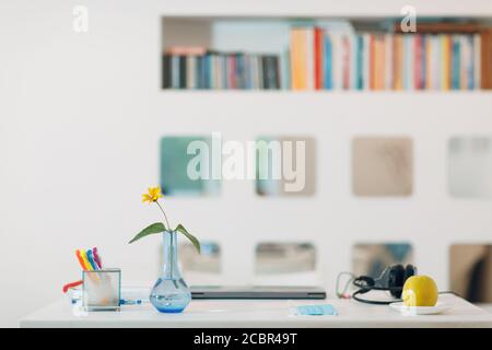 Moderne Haus Innenraum Arbeitsbereich mit einem silbernen Laptop, Kopfhörer, Vase, Blume, Apfel, Gesichtsmaske und Stifte auf dem Tisch mit weißen Wand Hintergrund und Stockfoto