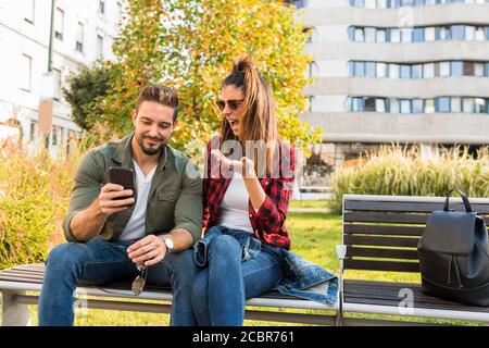 Ein Paar ist schockiert über Inhalte auf dem Bildschirm eines Telefons gesehen, während im Sommer auf einer Parkbank in der Stadt sitzen. Stockfoto