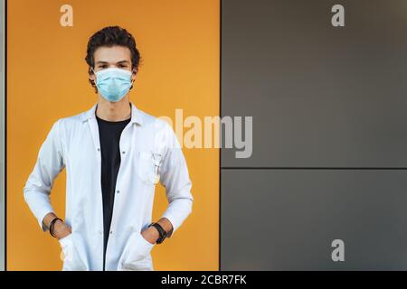 Portrait des Wissenschaftlers oder Arztes mit Maske und weißem Mantel Mit Händen in Taschen, die während des COVID-19-Ausbruchs gegen die Wand stehen Stockfoto