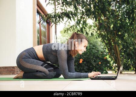 Volle Länge Seitenansicht der kaukasischen fit Frau kniet auf Übungsmatte während Sie sich online Tutorials auf dem Laptop auf der Terrasse ansehen Stockfoto