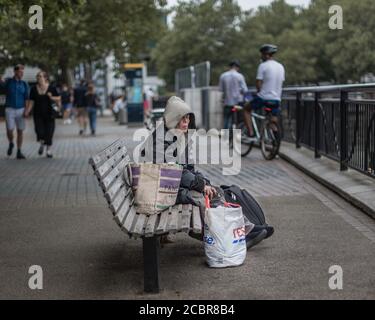 Als Touristen an der Straße vorbei gehen, sitzt eine Obdachlose auf einer Bank am Flussufer am Südufer der Themse in London. Stockfoto