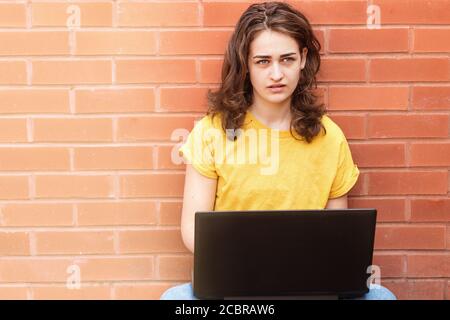 Traurige junge Frau in gelbem Hemd sitzt auf Flor, online studieren und arbeiten auf dem Laptop Stockfoto