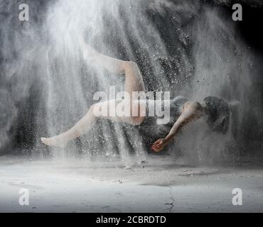 Schöne junge Frau in einem schwarzen Body mit einer Sportfigur schwebt auf einem schwarzen Hintergrund in einem Spray aus weißem Mehl, kreative Foto