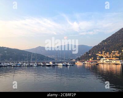 Comer See, Italien - Hügel, Berge und Blick aufs Wasser. Boote und Yachten in der Marina. Beruhigende Herbstszene, Sonnenuntergangslandschaft in sanften Farben Stockfoto