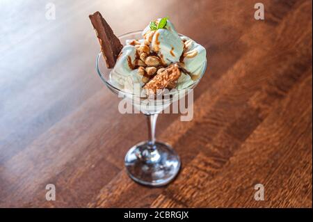 Dessert aus weißem Eis, Erdnüssen und einer Schokoladenriegel mit Minzblatt in einer Glasschale auf einem Holztisch Stockfoto