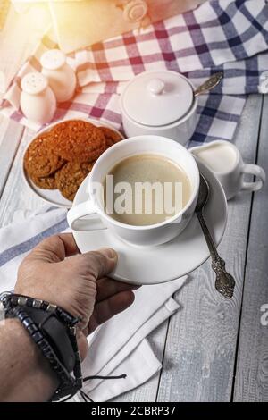 Mans Hand hält eine weiße Tasse Kaffee. Vor dem Hintergrund eines Holztisches mit Sahne, Zucker Schüssel und Haferflocken Cookies. Speicherplatz kopieren. Vertikale Sho Stockfoto