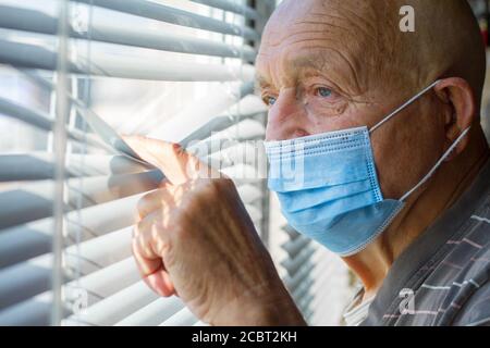 Sehr älterer Mann in blauer Schutzmaske auf seinem Gesicht schaut durch das Fenster, zu Hause bleiben Konzept Stockfoto