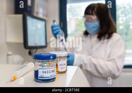 Entwicklung von Coronavirus-Impfstoffen. Glasfläschchen und Spritze auf dem automatisierten Laboranalysator. Wissenschaftlerin in Schutzbrille und Gesichtsmaske mit Pipette. Stockfoto