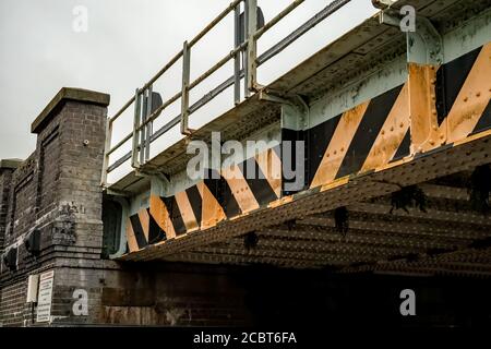 Nahaufnahme und Seitenansicht einer Eisenbahnbrücke aus Metall, mit Backsteinstützen und schwarzen und gelben Warnstreifen an der Außenseite. Stockfoto
