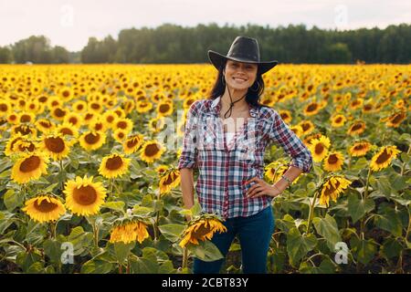 Junge Frau Agronomin trägt Cowboyhut, kariertes Hemd und Jeans auf Sonnenblumenfeld. Umfassendes Erntekonzept. Stockfoto