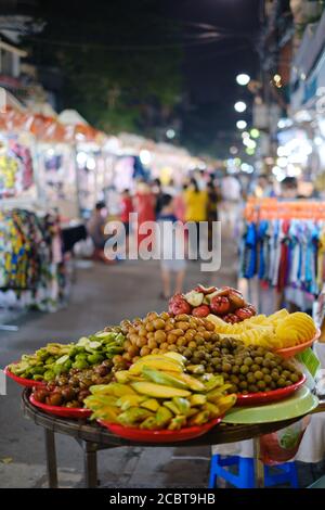 Frischer asiatischer Obstkorb in Hanoi Nachtmarkt, Vietnam verkauft. Obst im Fokus, verschwommener Hintergrund mit Menschen zu Fuß und Lichter. Stockfoto