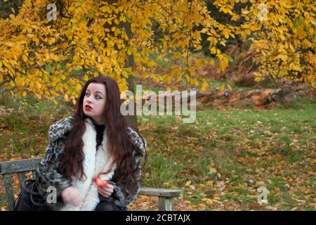 Eine kalte, besorgte junge Dame mit langen braunen Haaren saß an einem Herbsttag auf einer Bank unter einem goldgelben Baum Stockfoto
