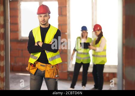 Männlicher Arbeiter auf einer Wohnbaustelle mit seinem Team Von Ingenieuren, die im Hintergrund arbeiten Stockfoto