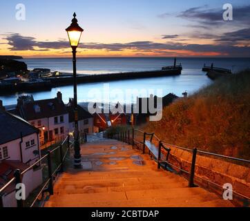 Bild zeigt die 199 Stufen zur abtei von whitby nach Sonnenuntergang mit einer Straßenlaterne und dem Hafen im Hintergrund. Whitby, North Yorkshire, England, Großbritannien. Stockfoto