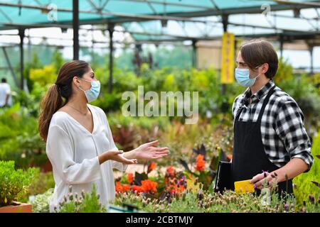 Junge Frau im Gespräch mit einem jungen männlichen Angestellten in einem Gartencenter, beide mit chirurgischen Masken. Gartenarbeit und Sicherheitskonzept. Stockfoto